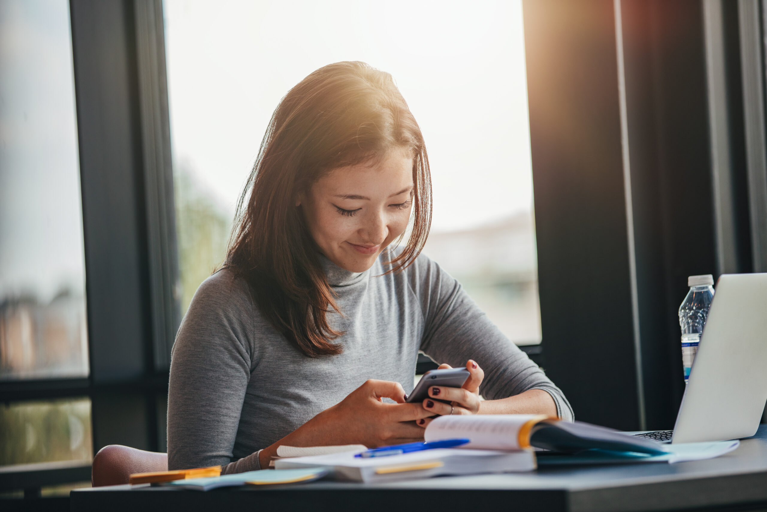 Shot of happy young female student sitting at table reading text message on her cell phone. Asian girl using mobile phone at college library.